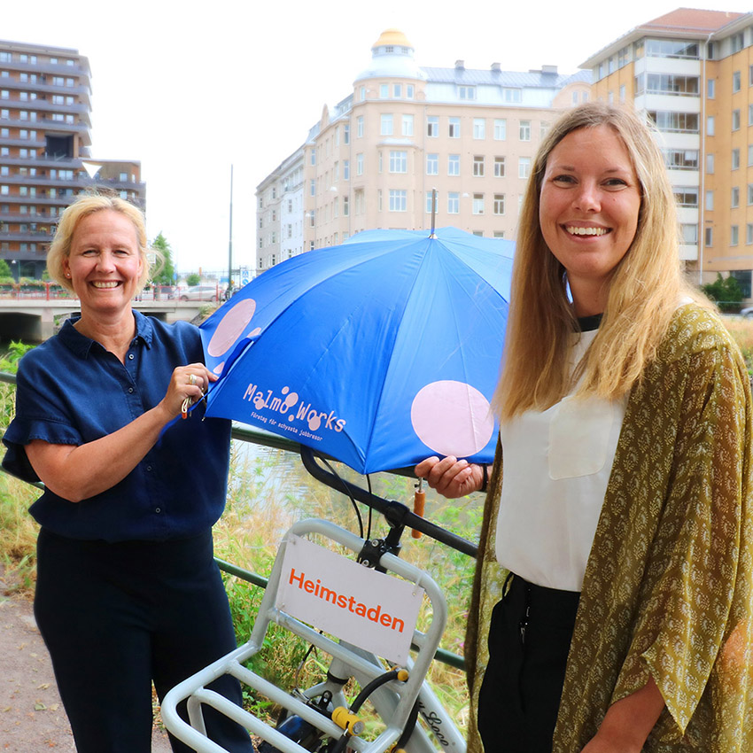 Två kvinnor står framför en cykel och håller ett blått paraply som det står Malmö Works på