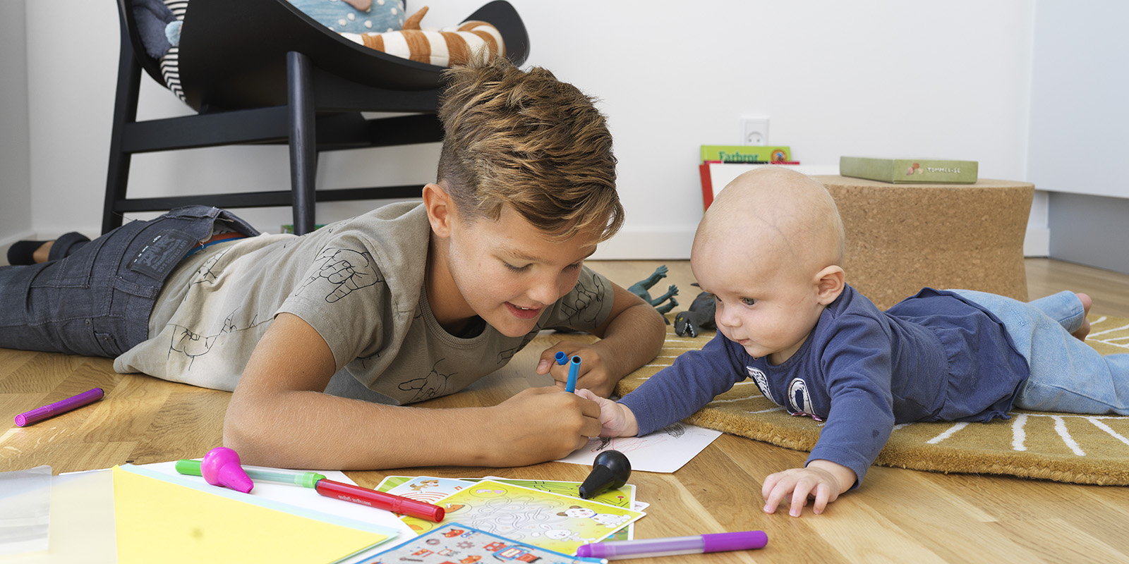 En ung pojke och en liten bebis ligger på golvet i en bostad och ritar på ett papper.