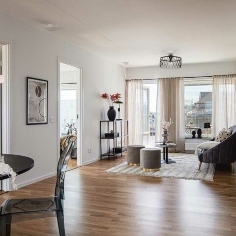 Ett vardagsrum med trägolv, stora fönsterpartier och möblerat med en beige tygsoffa och några sittpuffar i tyg.