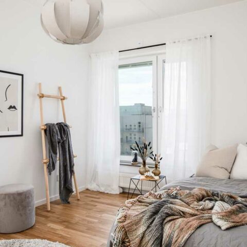 Ett vitt sovrum där man ser ett stort fönster med vita tunna gardiner, en bambustege som det hänger en morgonrock på. Sen ser man en del av sängen som är bäddad med ett grått överkast och en filt.