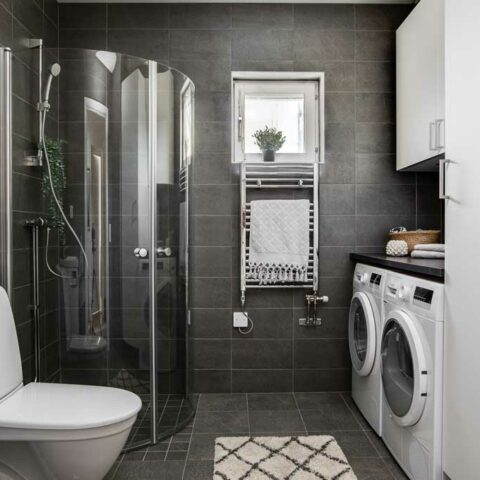 Ett mörkgrått badrum med en duschkabin i glas, tvättmaskin och en vit randig matta på golvet