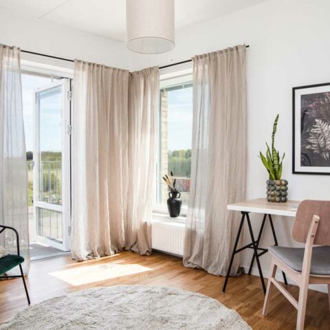 Ett ljust rum med tunna gardiner i fönstret. Mot väggen står ett litet vitt skrivbord och en trästol. På golvet ligger det en rund ljusgrå matta.