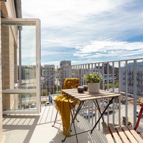 En balkong som ligger högt upp i ett hus. Ett litet cafébord och två trästolar står på balkongen.