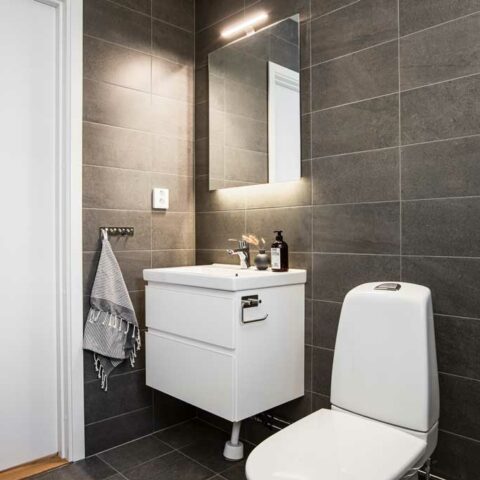 Ett mörkgrått badrum med fokus på en vit toalett, tvättställ och ett spegelskåp med belysning