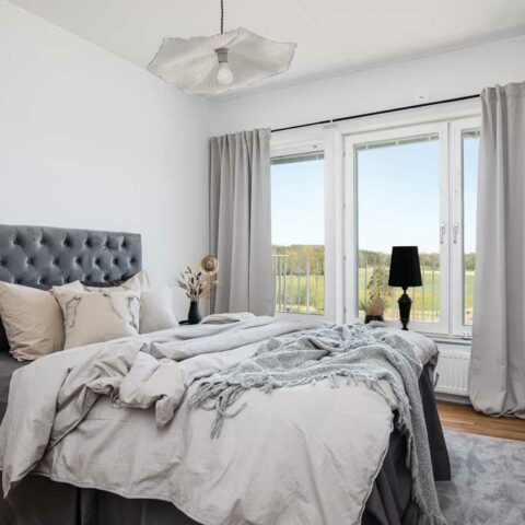 Ett ljust sovrum med en säng som har en stor grå sänggavel och bäddat i ljusgråa textilier