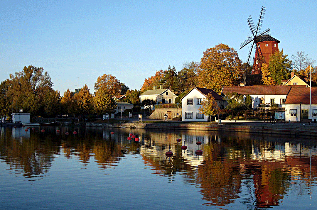 En bild tagen på hösten där man ser en rödbrun mölla och gamla vita tegelbyggnader vid vattnet.