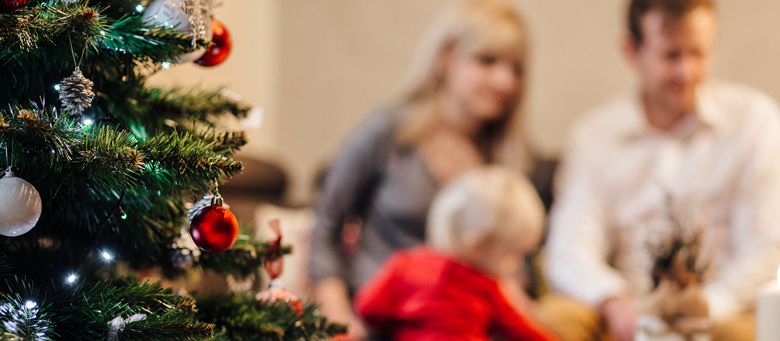 En grön julgran med fina dekorationer och suddigt i bakgrunden ser man en kvinna, man och ett barn i röd tröja