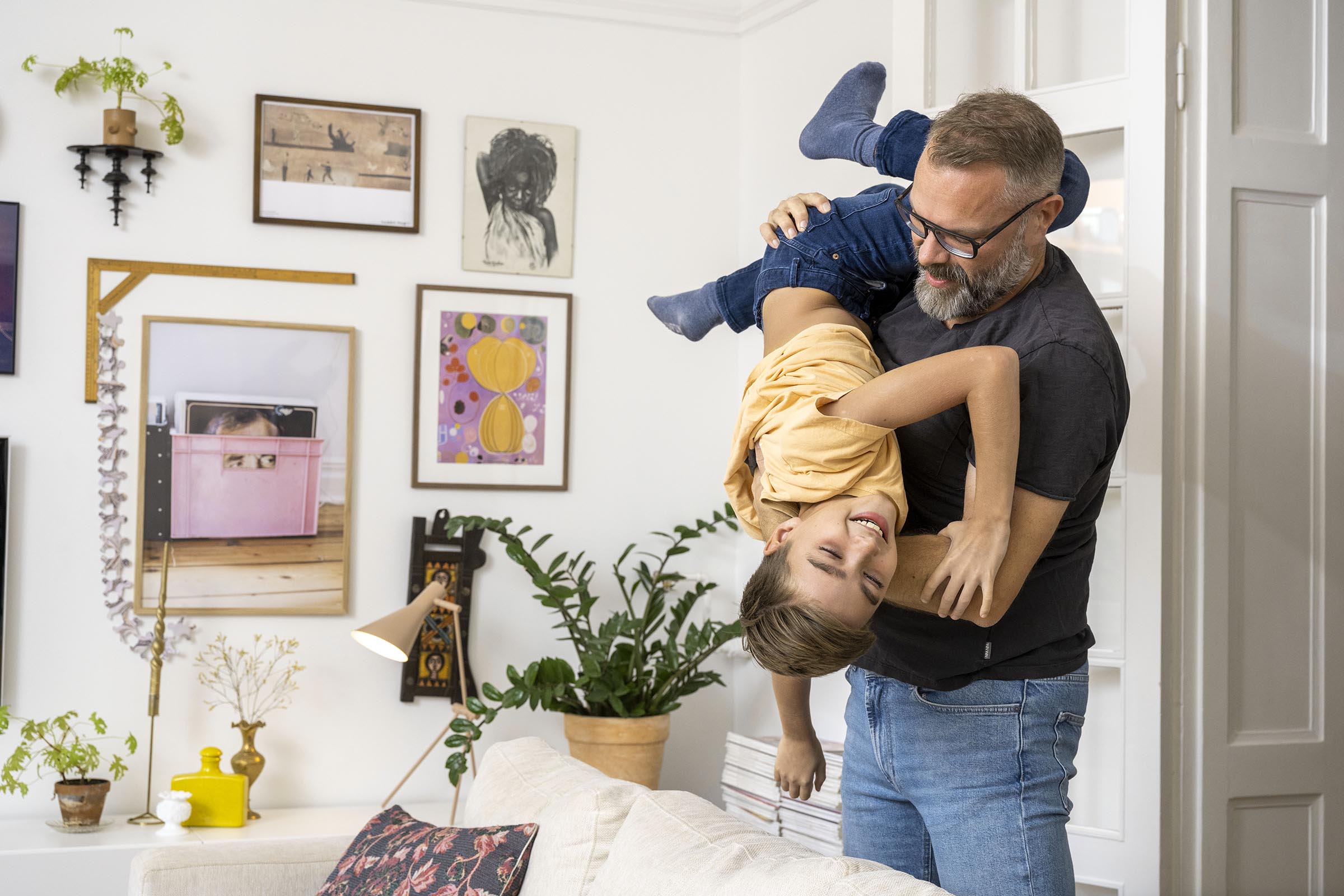 En man med skägg, jeans och grå tröja busar med en pojke med gul tröja i ett vardagsrum