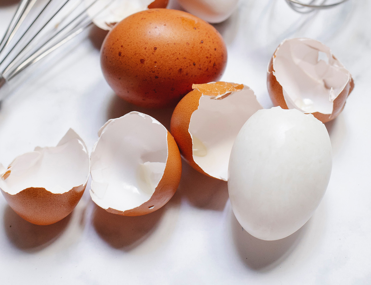 En vit duk med ett helt brunt ägg på och fyra äggskal. Det ligger även en metallvisp uppe i vänstra hörnet.