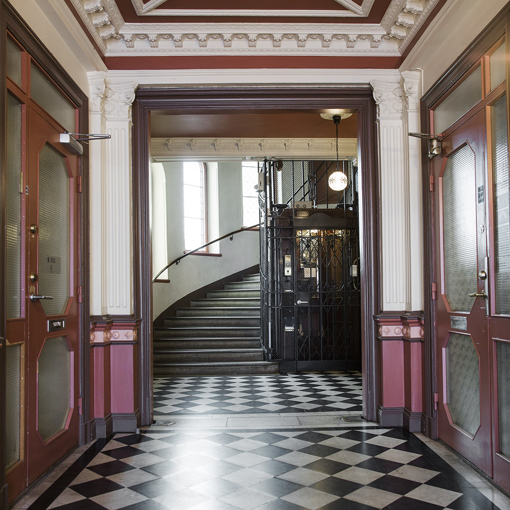 Ett trapphus med svartvitt klinkergolv och en stor mörkbrun trappa