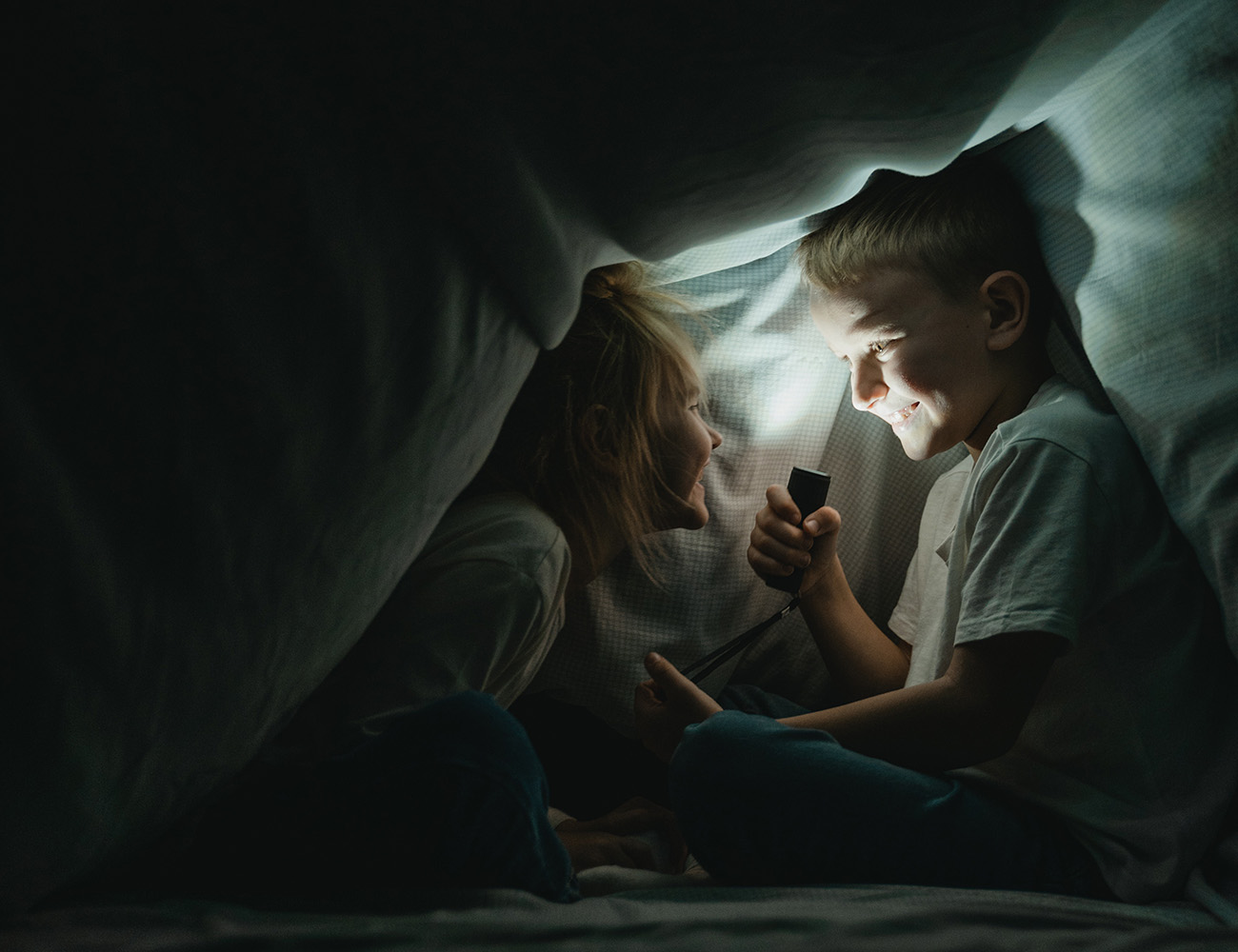En pojke och en flicka är under ett stort täcke och lyser med ficklampan i ansiktet