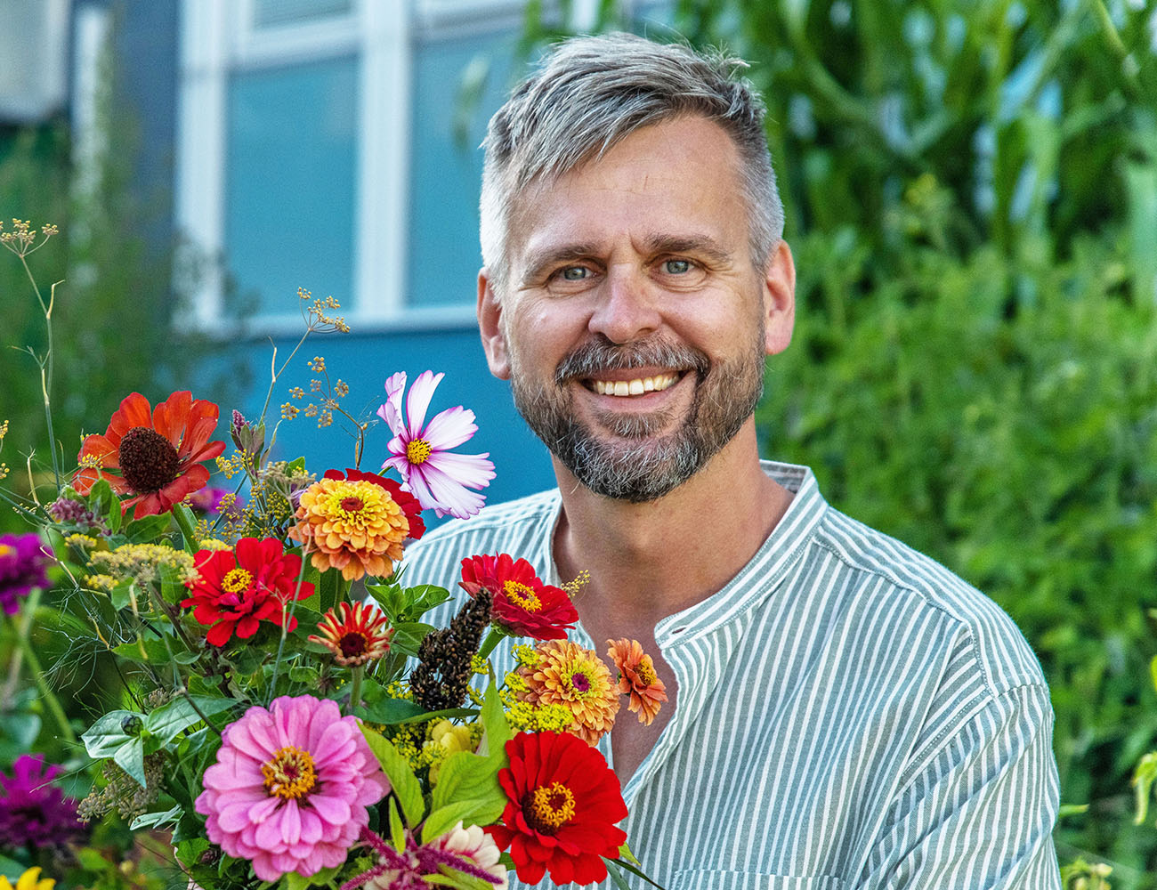 En man med grått hår och skägg och en randig skjorta har en stor bukett blommor i rosa, rött och lila framför sig. Bakom ser man en blå husfasad och lite träd.
