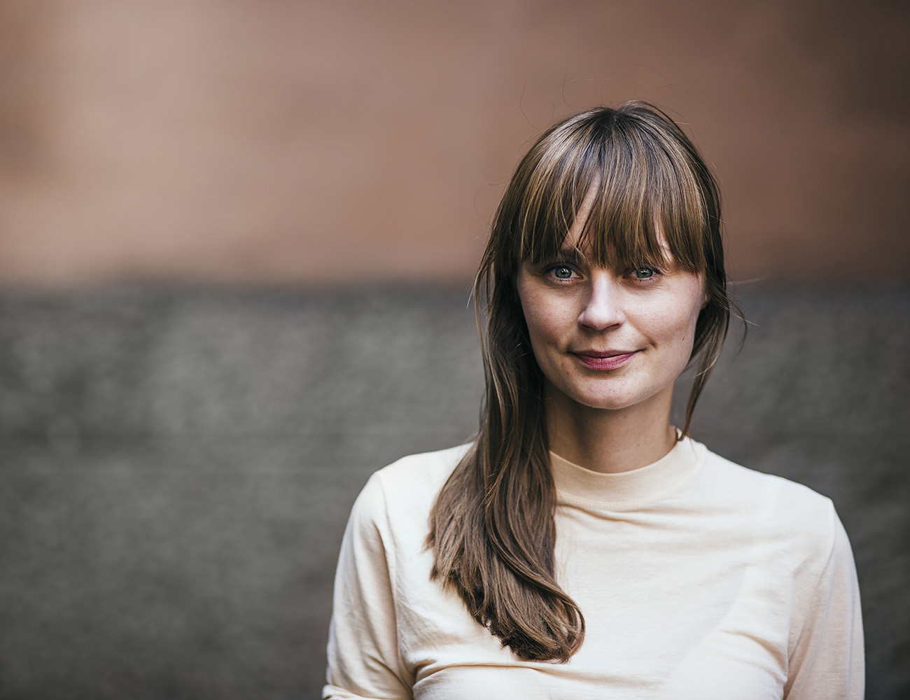 En mörkhårig kvinna med långt hår och lugg och en ljus polotröja ler och tittar rakt in i kameran.