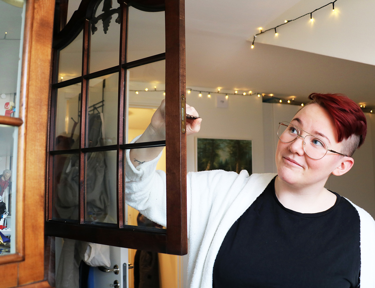 En tjej med kort rött hår, svart tröja och en vit kofta håller i handtaget på en vitrinskåpsdörr som är i trä.