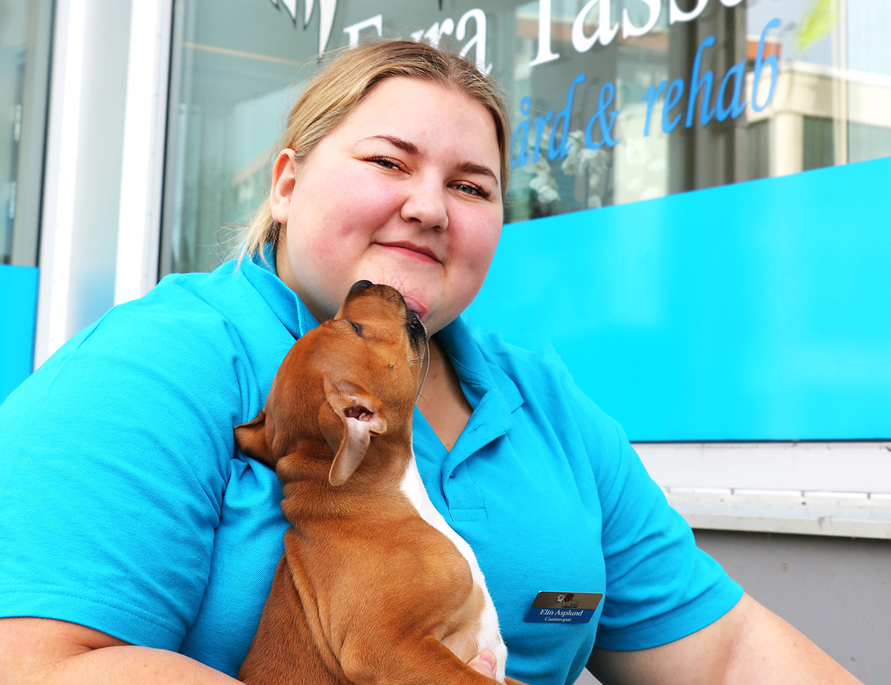 En ljushårig tjej med en knallblå pikétröja sitter utanför en hundbutik som heter Hyra tassar. I hennes famn har hon en liten brun och vit hundvalp som slickar henne i ansiktet.