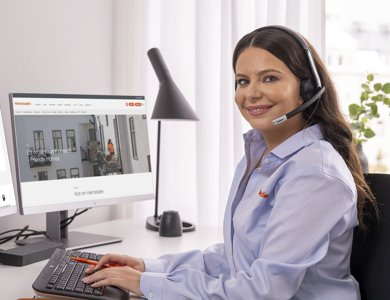 En mörkhårig kvinna med ljusblå skjorta sitter med sitt telefon headset framför datorn