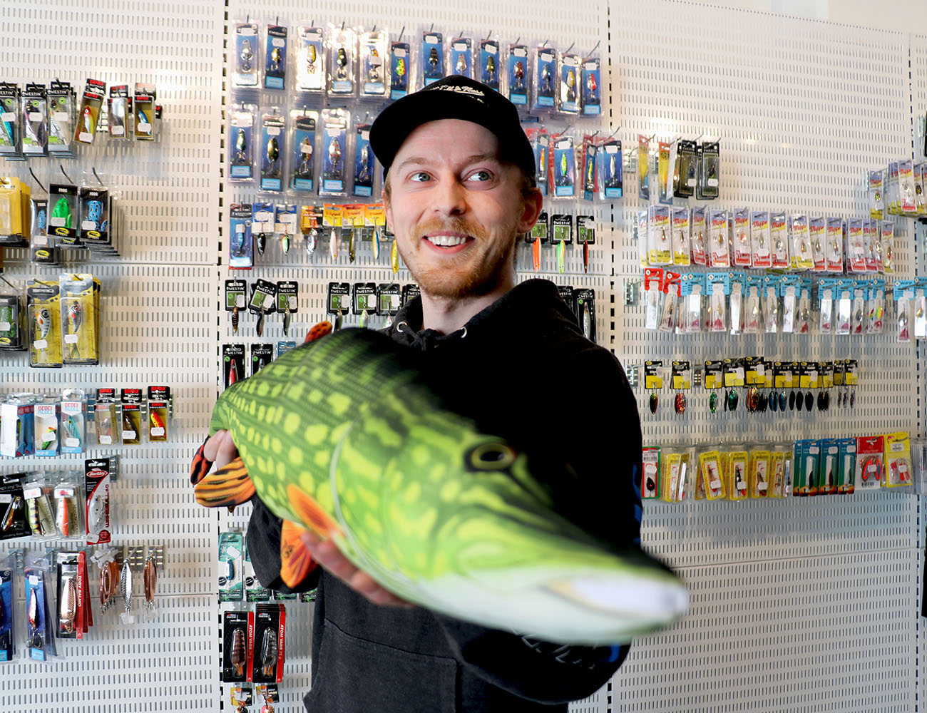 En kille med svart hoodtröja och svart keps står i en fiskebutik med en jättestor grön fisk i handen