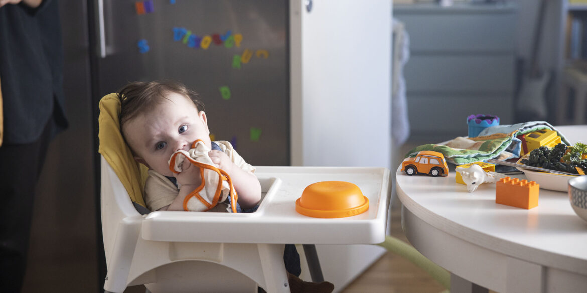 En bebis sitter i en barnstol i plast och tuggar lite på en tyghaklapp. Framför hen så är det lite färgglada leksaker och matskålar.