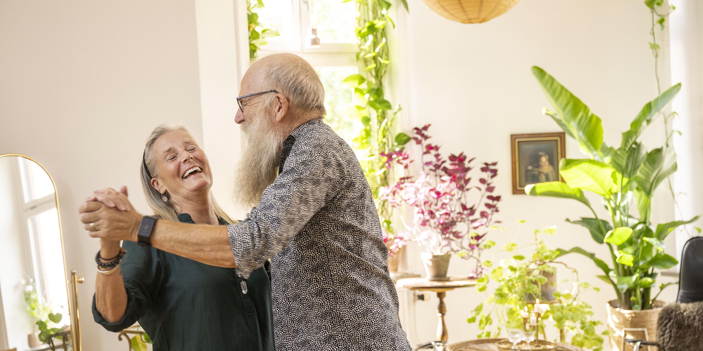 En äldre man och kvinna dansar i sitt vardagsrum med massa gröna växter och skrattar samtidigt.