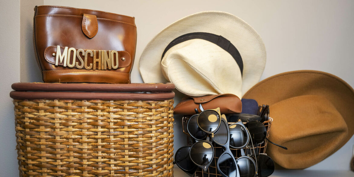 En hylla där det står en rottingkorg med lock, en vit och en brun hatt samt en korg med massa solglasögon i
