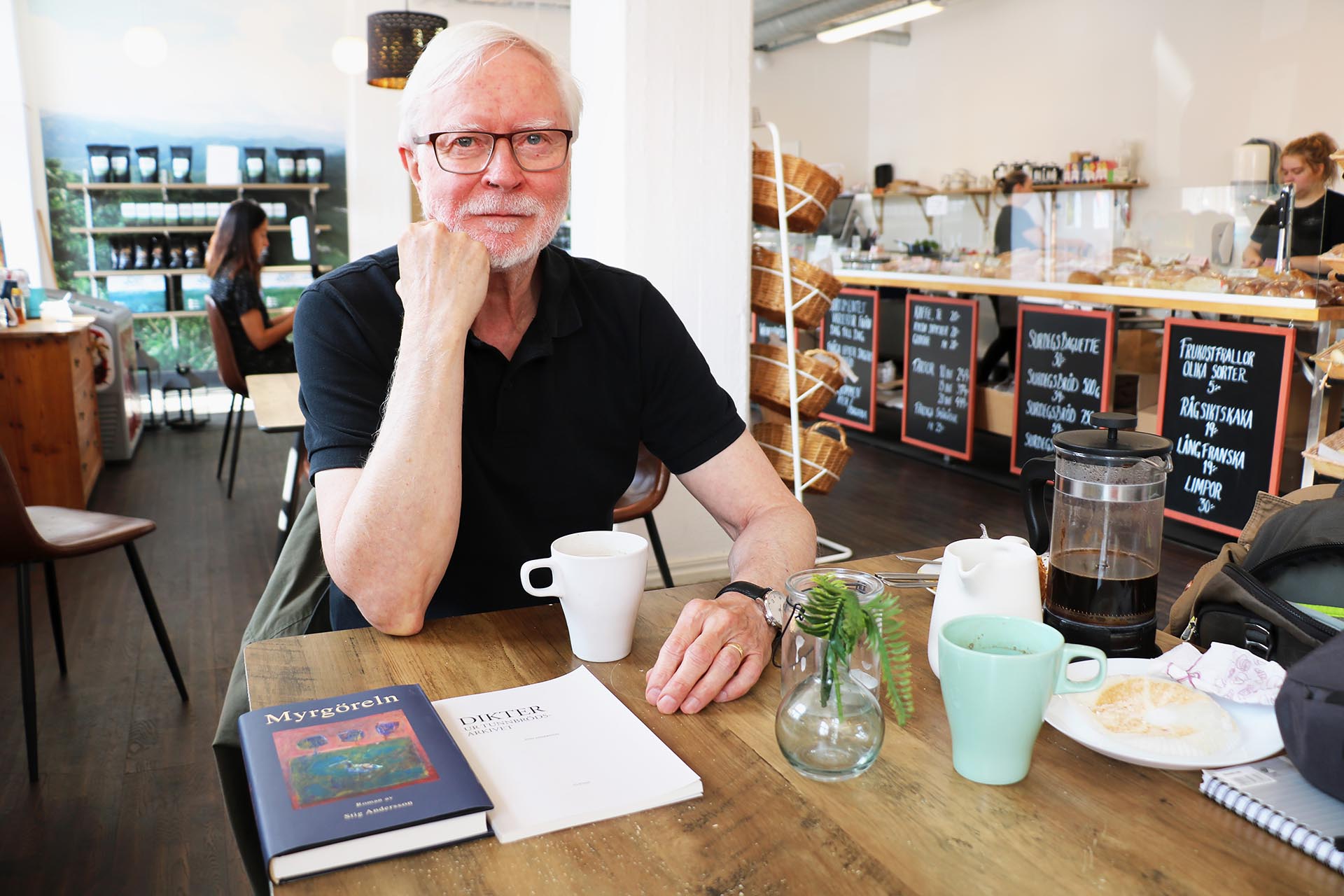 En man med svart tröja och grått hår sitter på ett fik med en kaffekopp och böcker framför sig