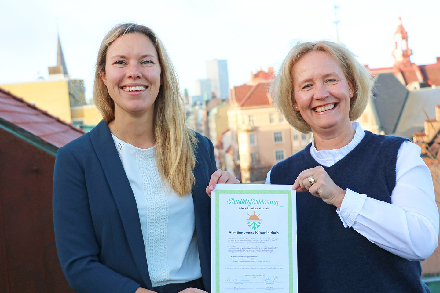 Två ljushåriga kvinnor står på en balkong utomhus hållandes i ett diplom. I bakgrunden ser man en storstadsvy.
