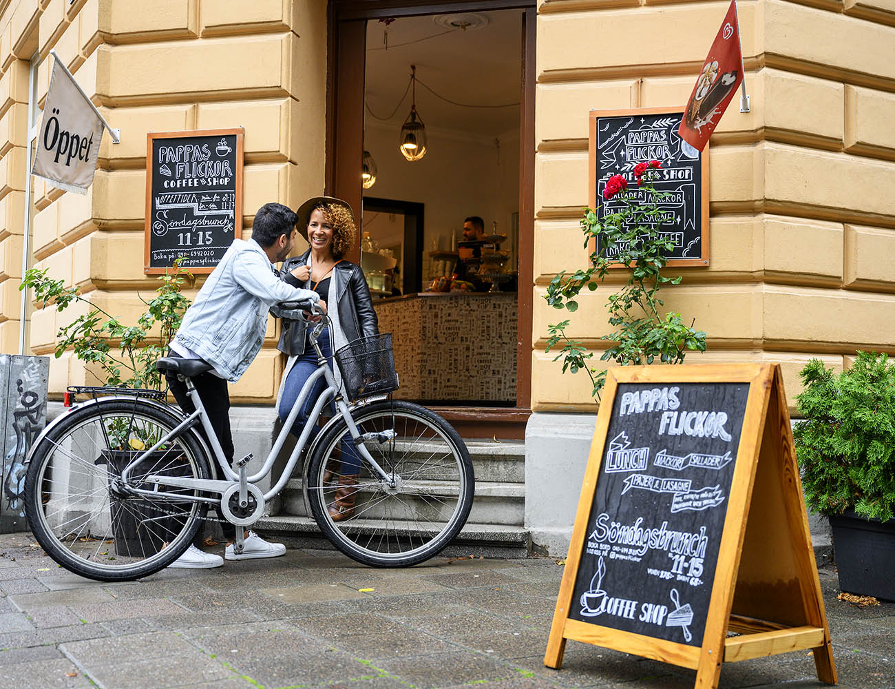 En tjej och en kille står utanför ett fik och pratar. Killen håller i en cykel och ute på gatan ser man en trottoartalare och skyltar där man kan se vad fiket erbjuder för meny.