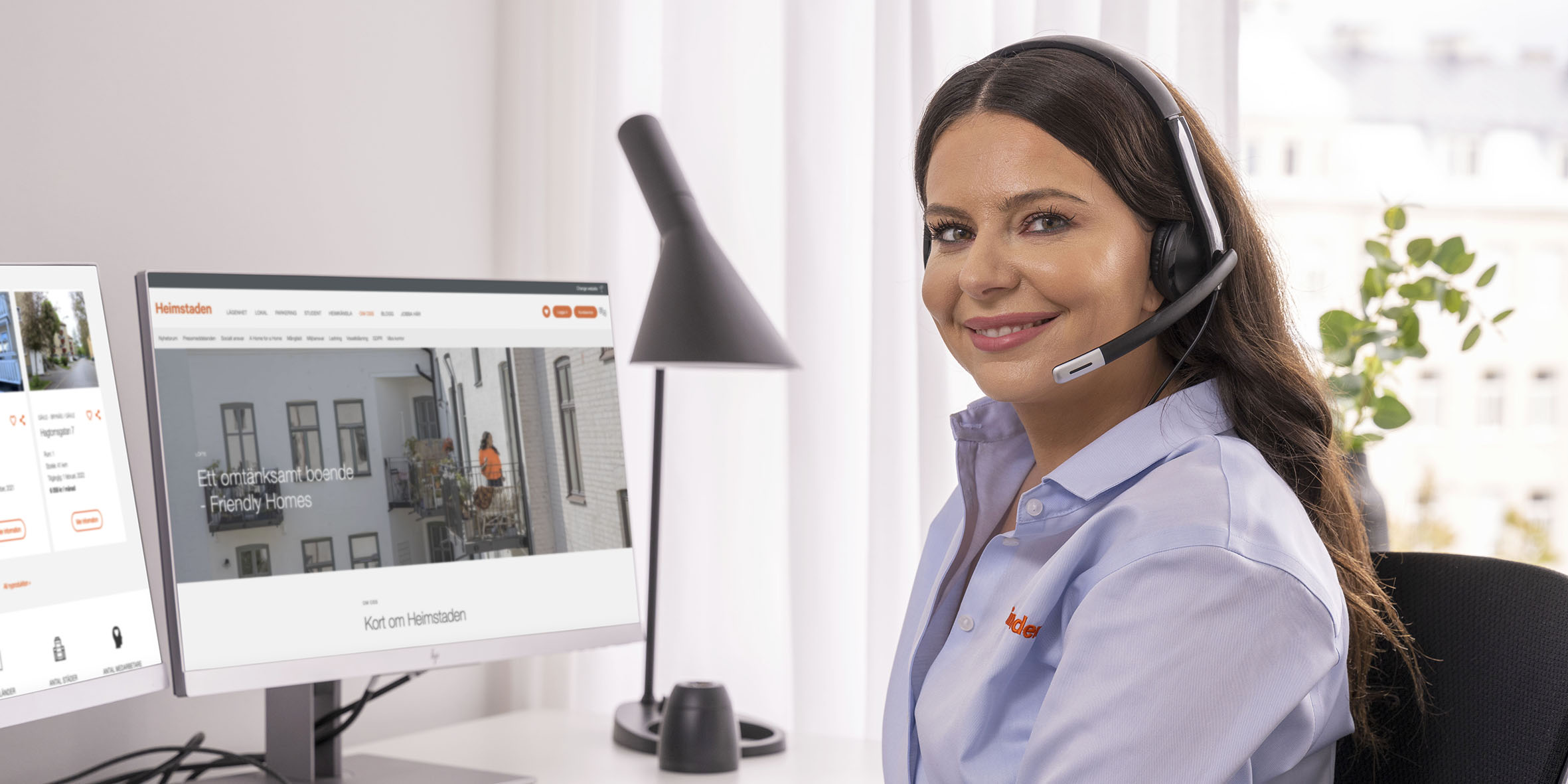 En kvinna med mörkt hår sitter med sitt headset framför datorn och ler in i kameran