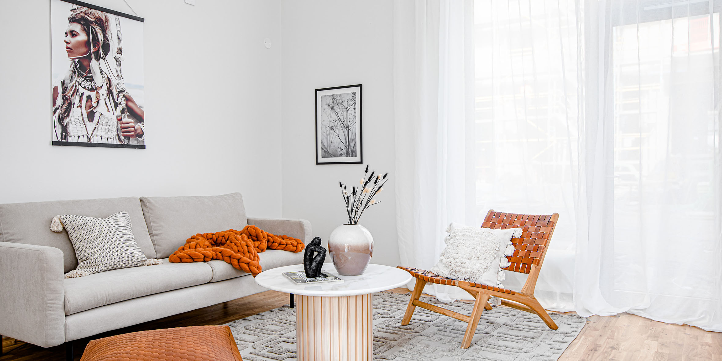 Ett ljust och vitt vardagsrum med en beige soffa och en skinnfåtölj. En orange filt och en stor tavla med ett motiv av en kvinna syns också.