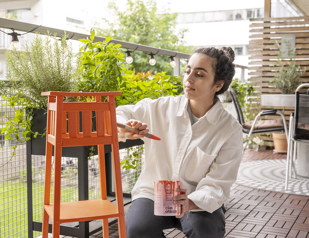 En ung tjej med en vit skjorta sitter på sin balkong och målar en växtmöbel i en orange färg.
