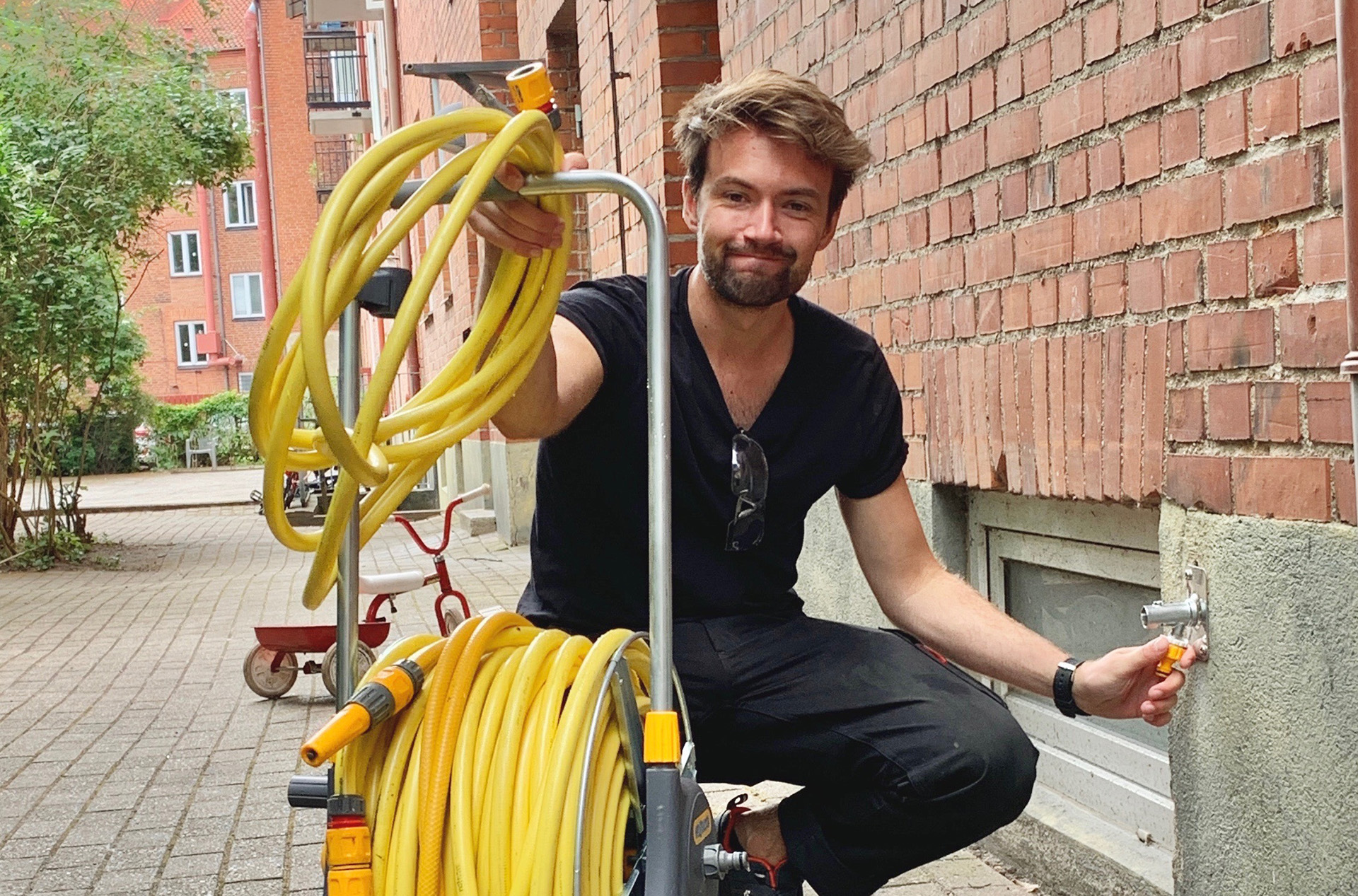 En man med svarta kläder står på huk i en innergård och håller i en gul vattenslanganordning.