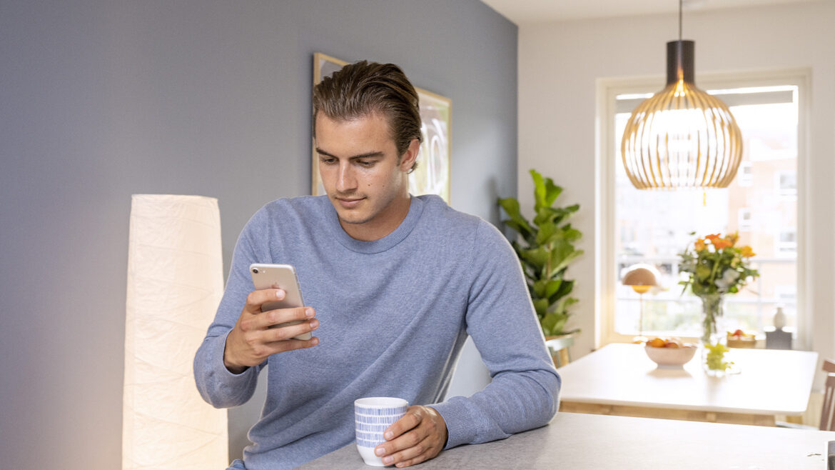 En ung kille med blå tröja sitter i köket och har en kaffekopp i ena handen och en mobil i andra.