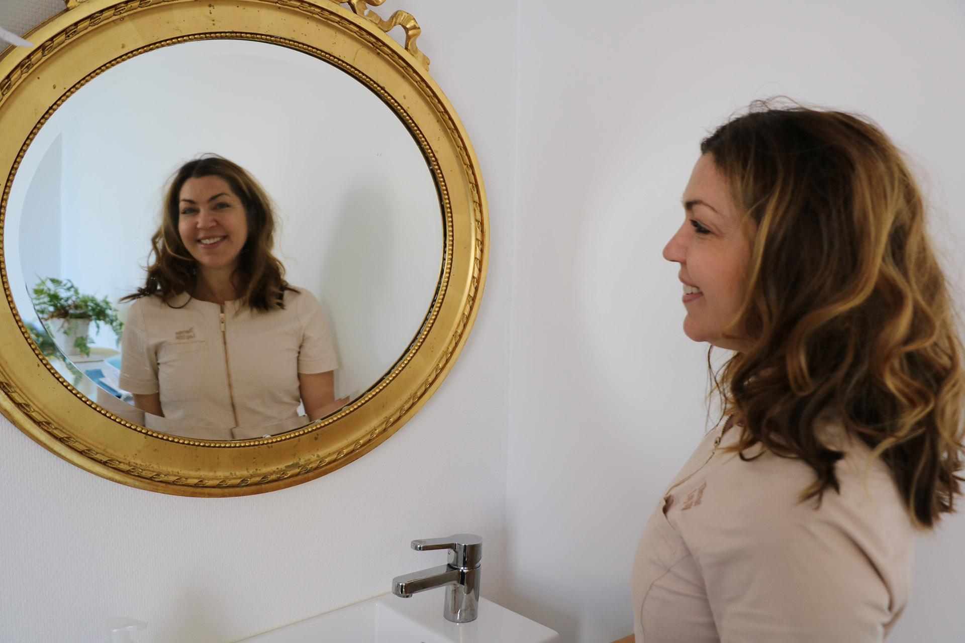 En kvinna med lockigt mörkt hår står framför en rund guldspegel och ler.
