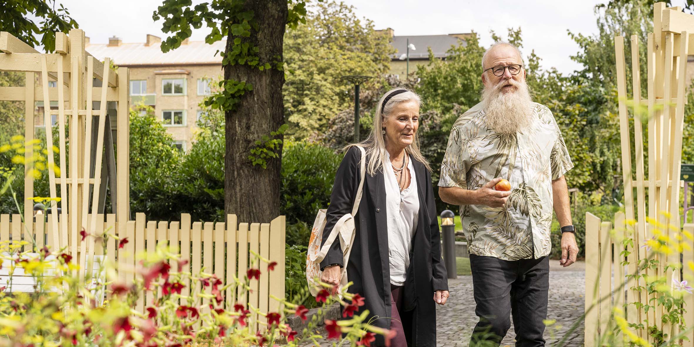 En äldre kvinna och man i stort skägg och glasögon går i en grön park med färgglada blommor och gula staket.