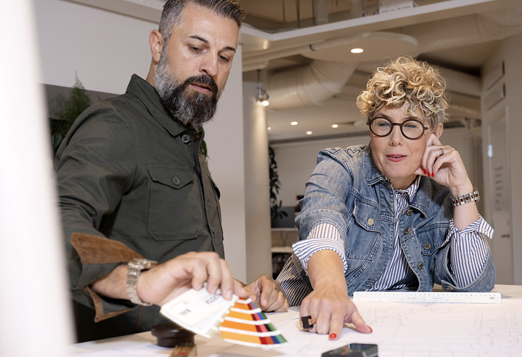 En man i mörkt skägg och en kvinna med kort lockigt hår och glasögon står vid ett bord och tittar på färgprover och ritningar.