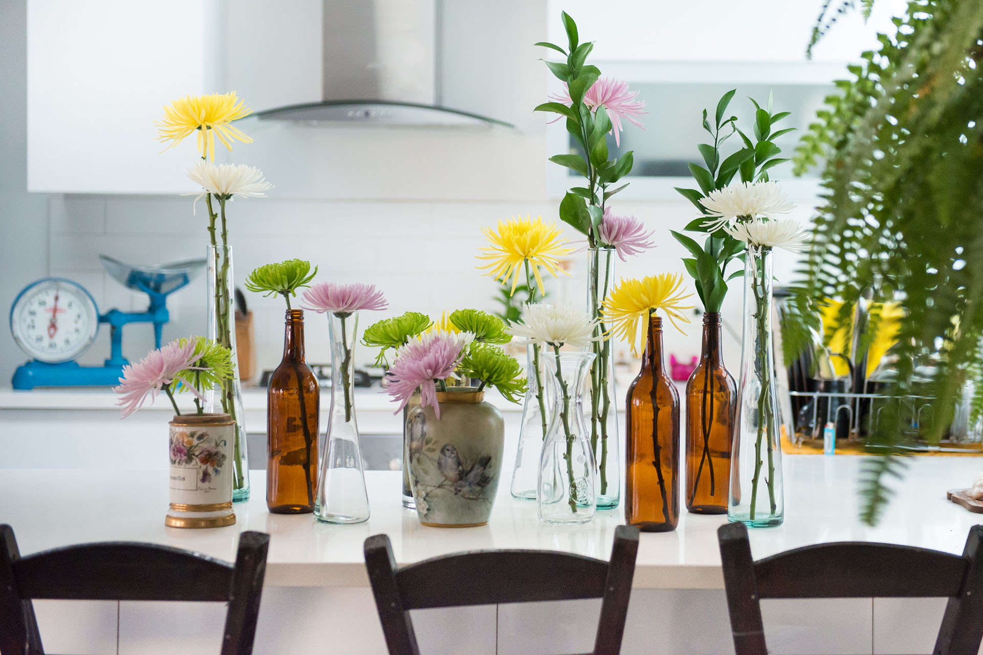 Ett bord med en vit duk på som det står flaskor och vaser på med blommor i. Vid bordet står det tre svarta köksstolar. Bakom bordet ser man en köksbänk som det står en blå våg på.