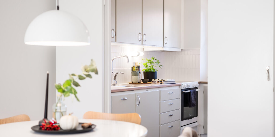 Ett ljusgrått kök i 30-tals stil där man ser gröna örter och skärbrädor på diskbänken. I hörnet ser man en del av ett matbord och en vit taklampa