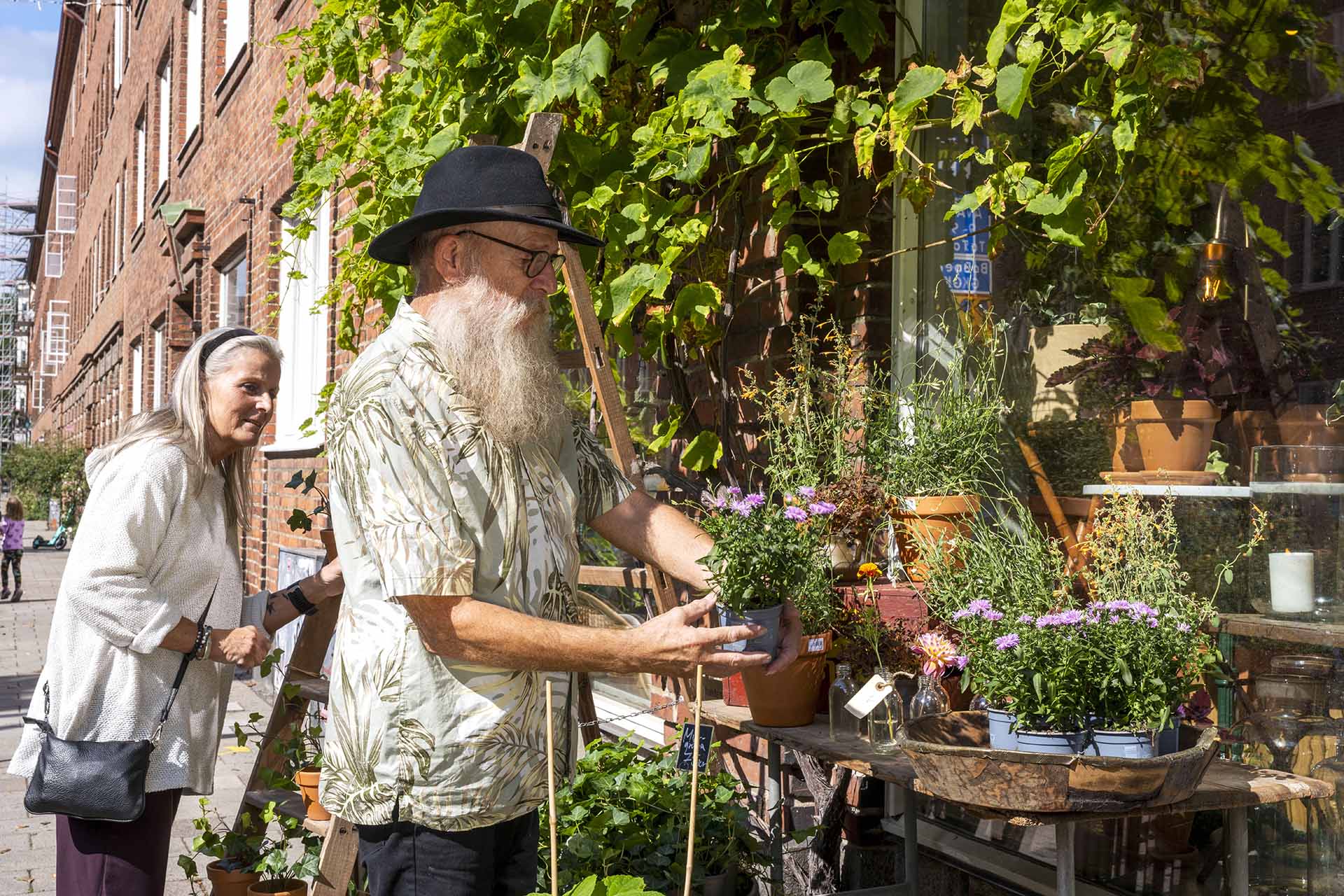 En man med vitt skägg och svart hatt står utanför en blomsterbutik och tittar på en krukväxt. En kvinna med grått hår står vid sidan om och tittar nyfiket.