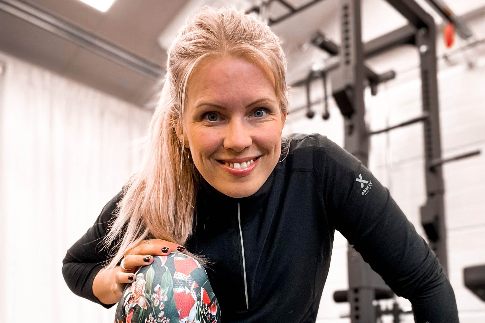 En blond tjej med uppsatt hår sitter på huk i ett gym med träningskläder på sig. Bakom henne ser man träningsmaskiner.