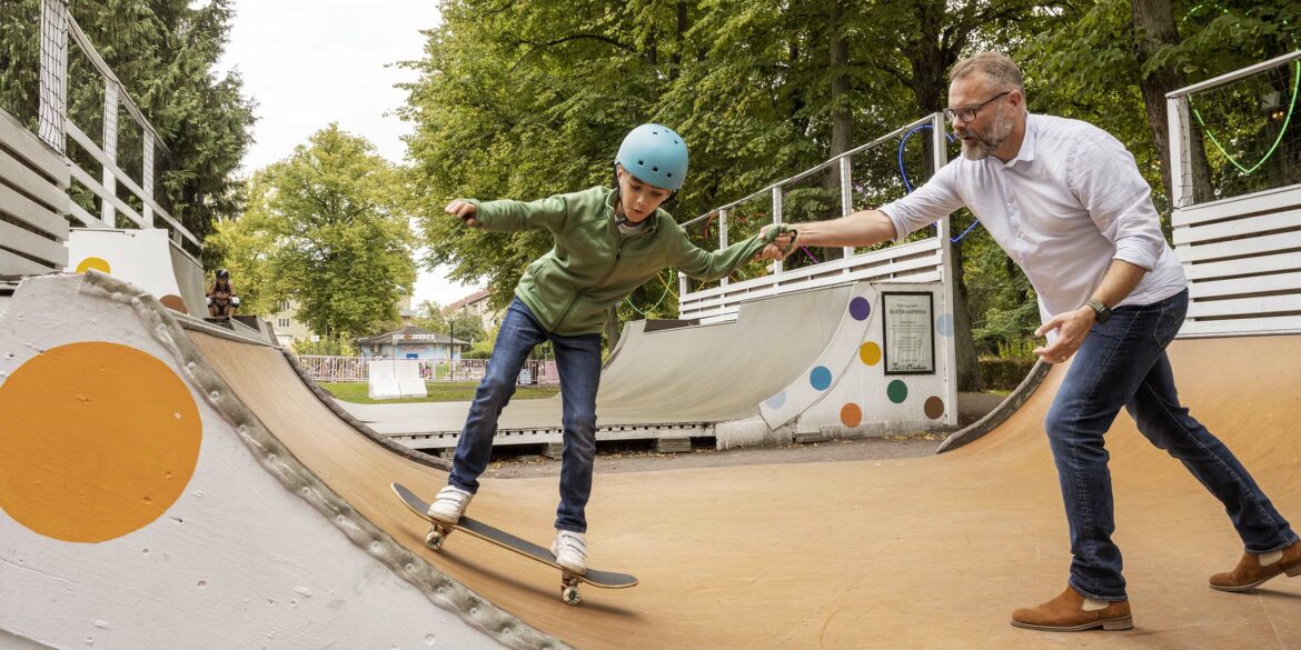 En man i skjorta och jeans hjälper en pojke i en skateboardramp som åker på en bräda med en blå hjälm.