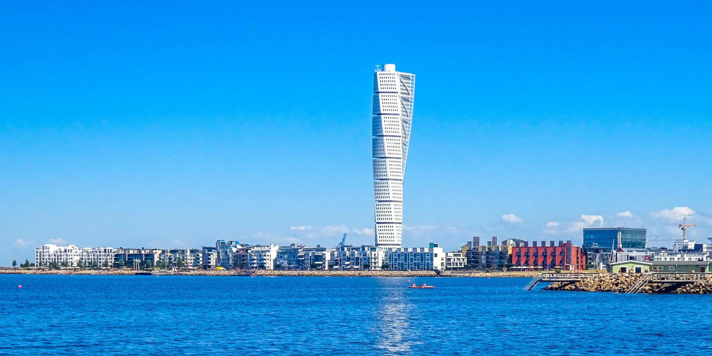 En stor vit skyskrapa som ser tvistad ut, Turning torso i Malmö. Bakom ser man en knallblå himmel och blått hav framför. Man ser låga hus precis vid strandkanten.