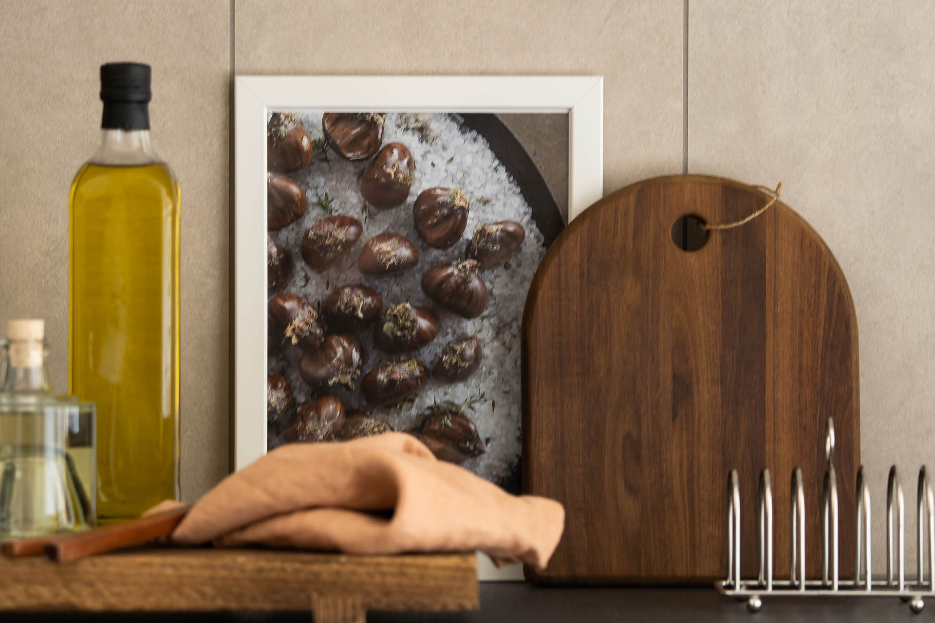 En närbild på en köksbänk där det står en oljeflaska, skärbräda i trä, en liten tavla med fikonmotiv och en rostfri toastbröd hållare.
