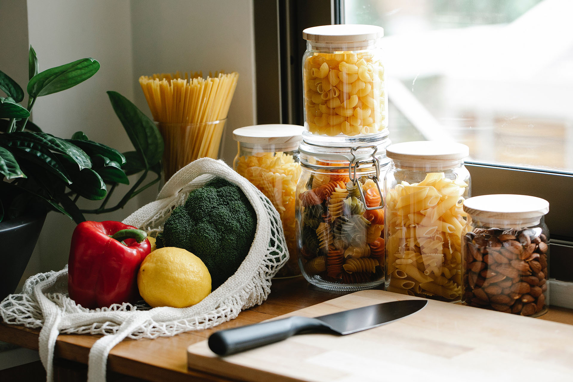 En köksbänk som det står olika glasburkar på med pasta i olika färger i. Framför ligger en skärbräda i trä med en kniv på och sen vit nätpåse med en citron och röd och grön paprika i