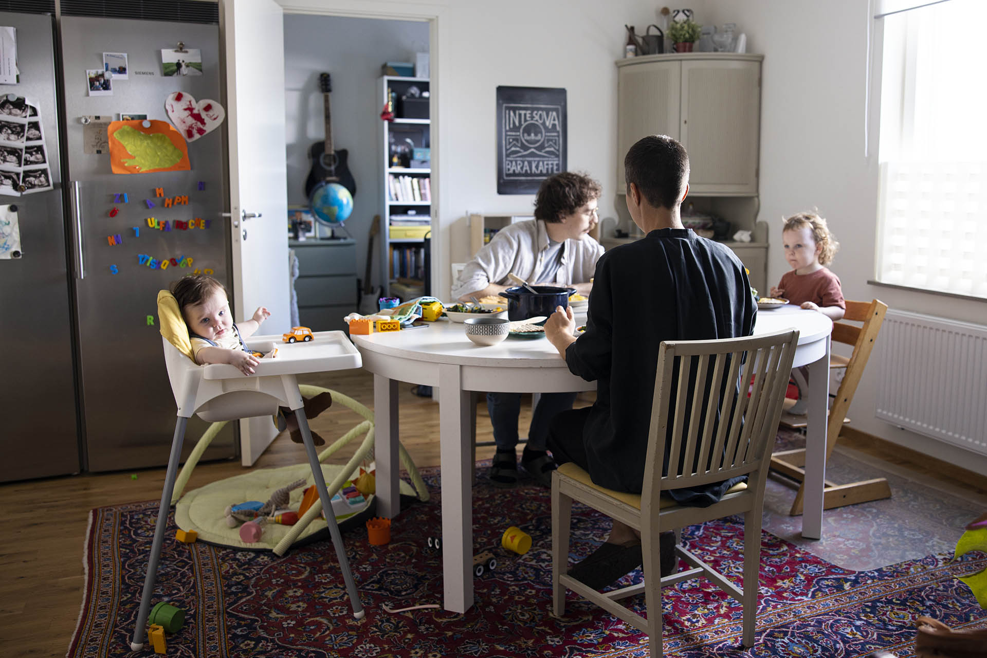 En familj som sitter vid ett matbord i sitt kök och äter. Under bordet ligger det en stor persisk matta i rött och blått. På det rostfria kylskåpet ser man massa magneter och barnteckningar.