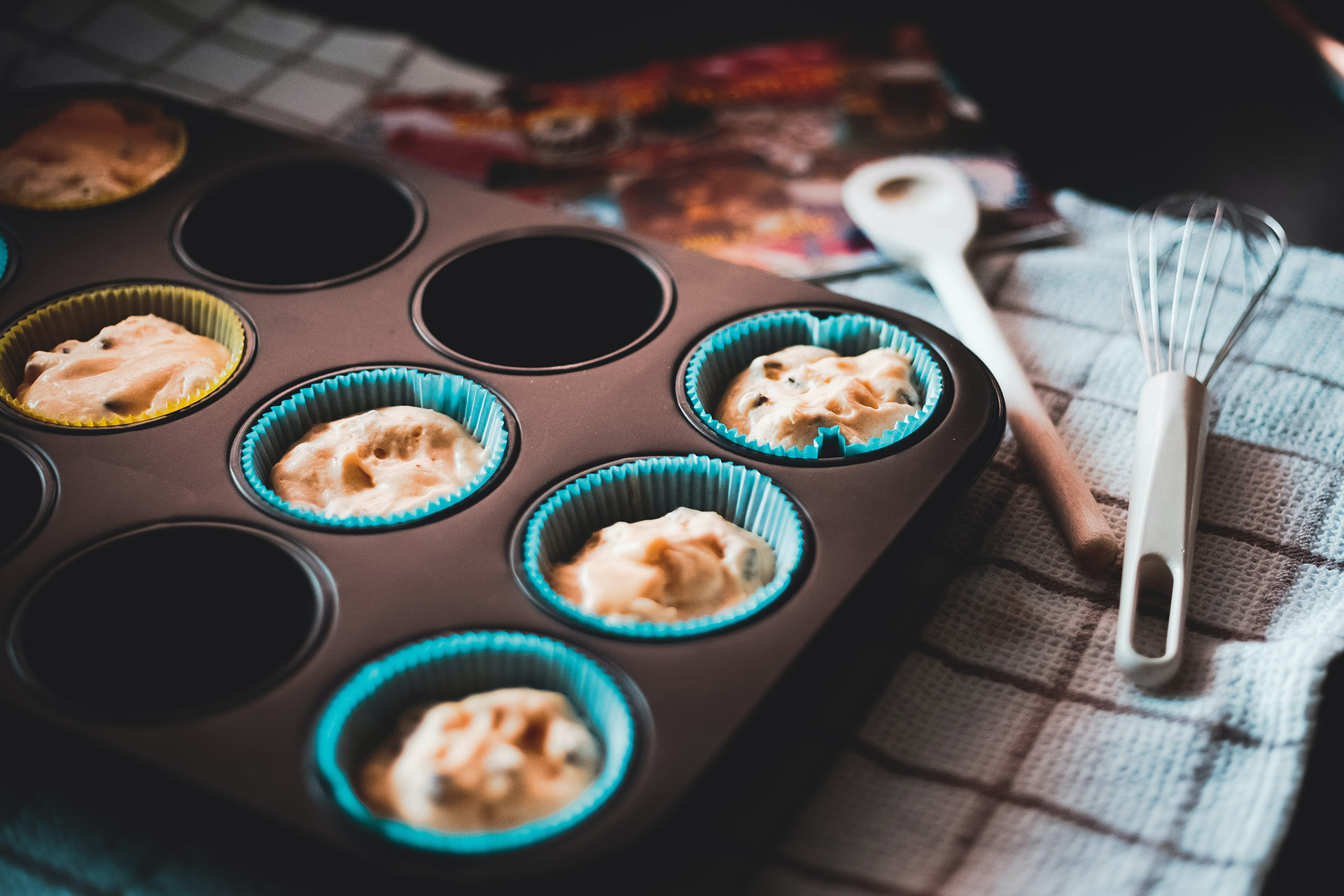 En svart muffinsform som ligger på en vit och blå rutig duk. I formen ligger det muffinsformar och deg som är redo att åka in i ugnen och bakas. På duken ligger även en träslev och en tidning.