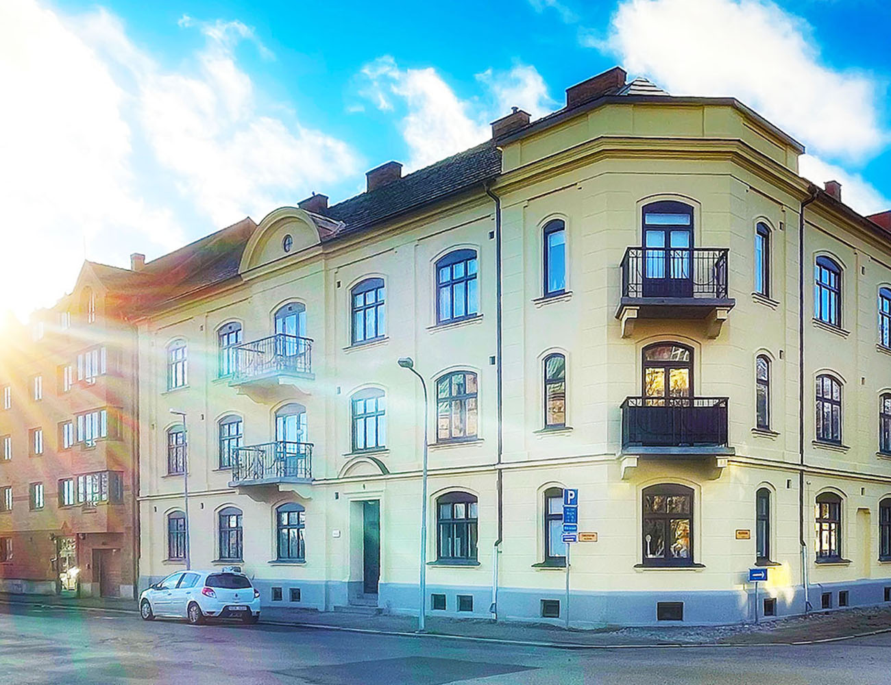 Ett trevåningshus i en ljusgul putsad färg. Man ser två hörnbalkonger samt en vit bil som står parkerad på gatan.