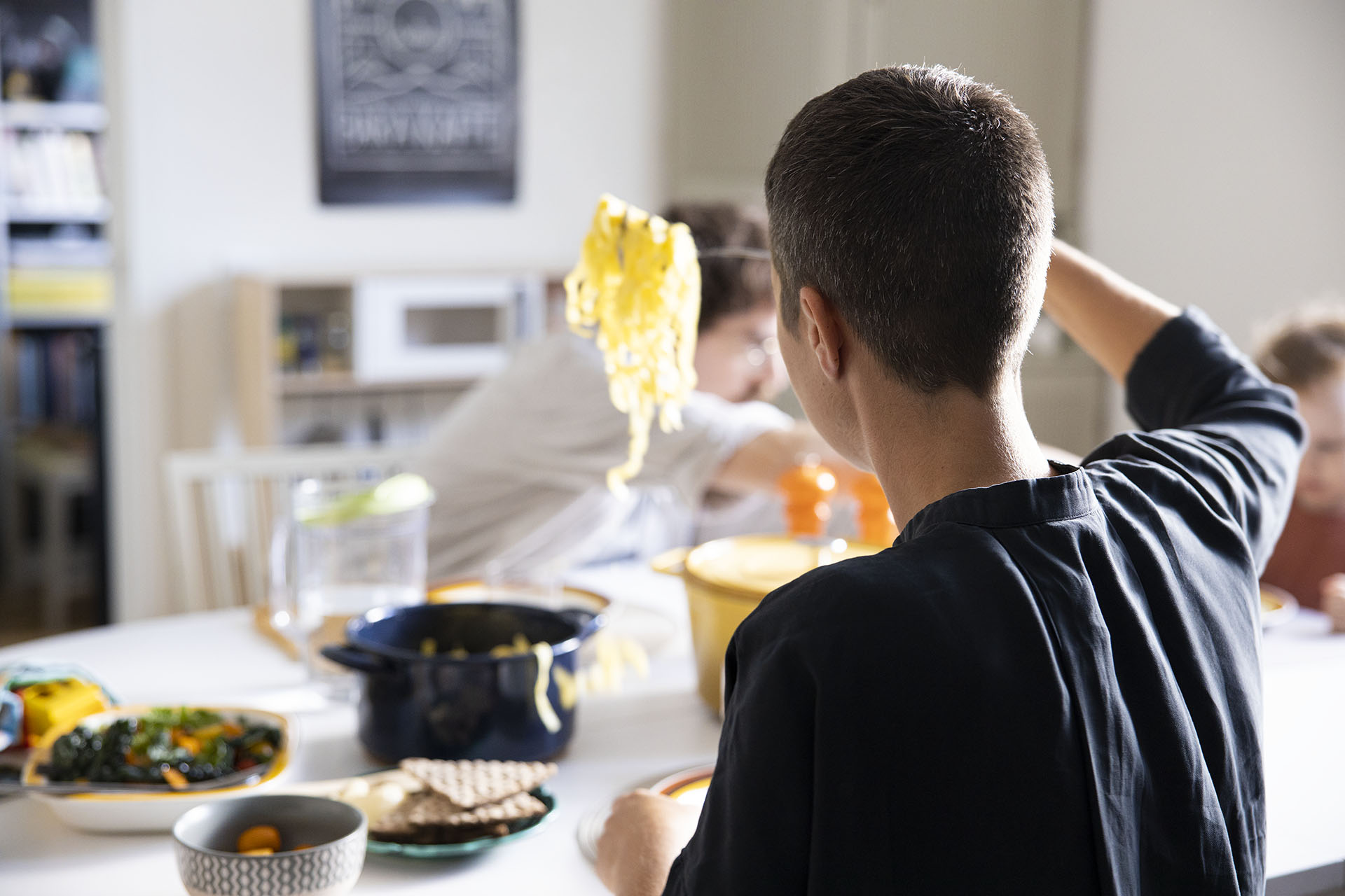 En kvinna med kort hår sitter vid ett köksbord och håller upp en slev med spaghetti ovanför hennes tallrik. Hon sitter med ryggen mot kameran och ha en gråsvart tröja på sig.