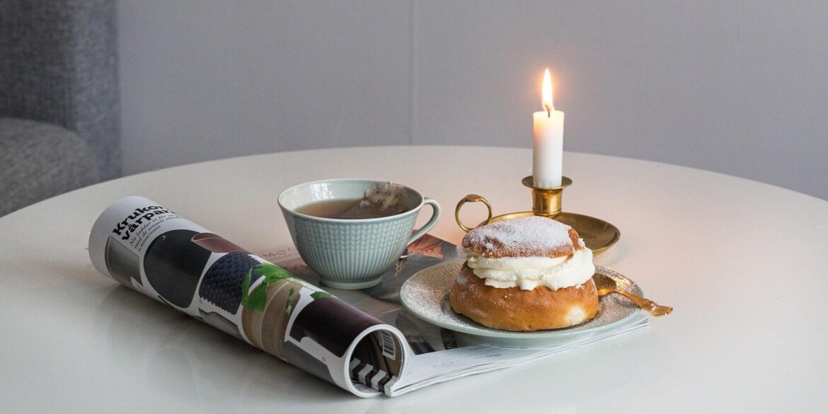 Ett vitt köksbord som det står ett fat med en semla på, en ljusblå kaffekopp och ett stearinljus i en mässingsstake. Det ligger även en ihoprullad tidning under kaffekoppen.