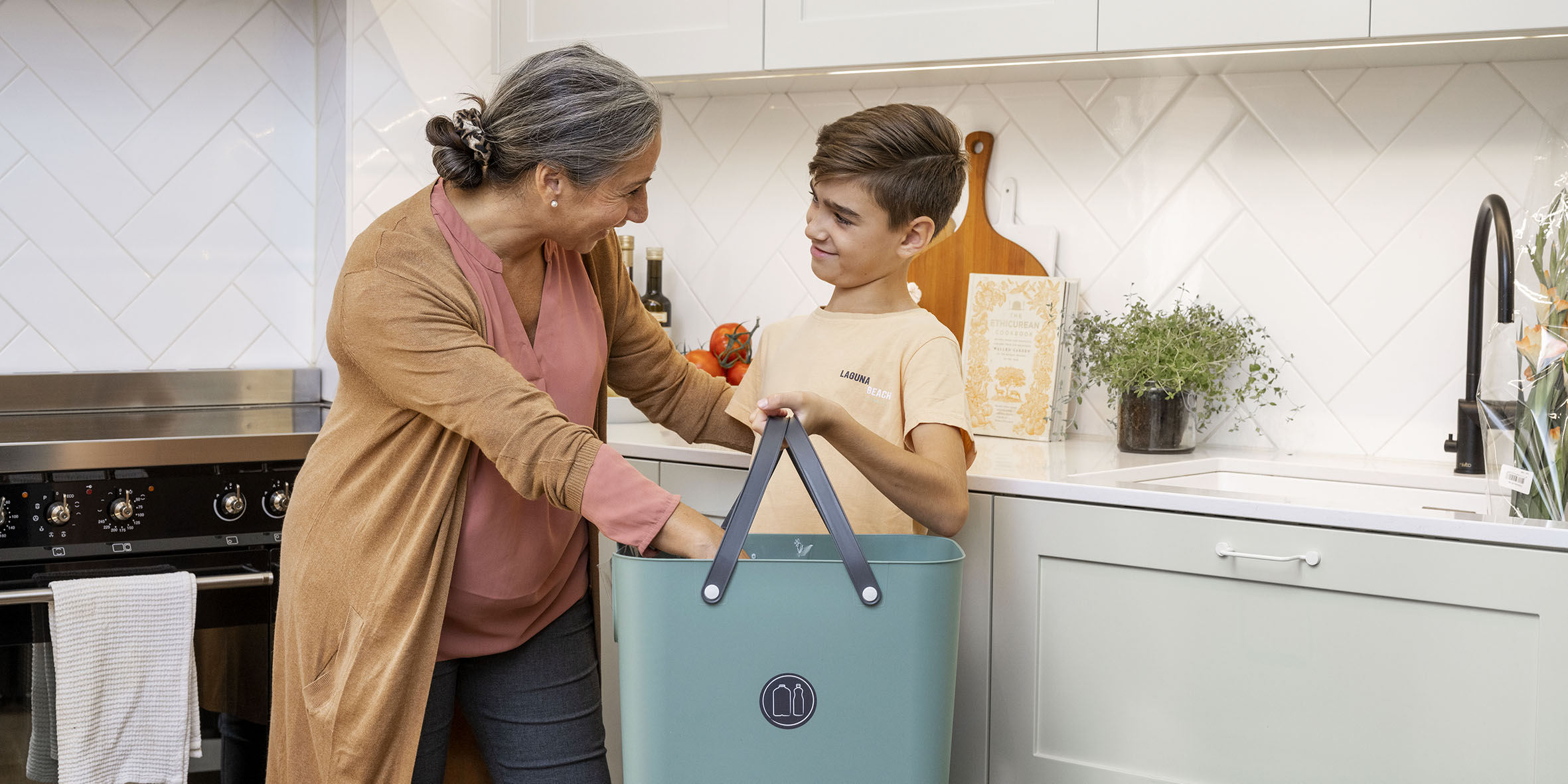 En kvinna med gråsvart hår och en rosa tröja står i ett kök tillsammans med en pojke i gul tröja. De håller med varsin hand i en blå plastlåda med handtag. Bakom ser man en diskbänk med några träskärbrädor och en kokbok.