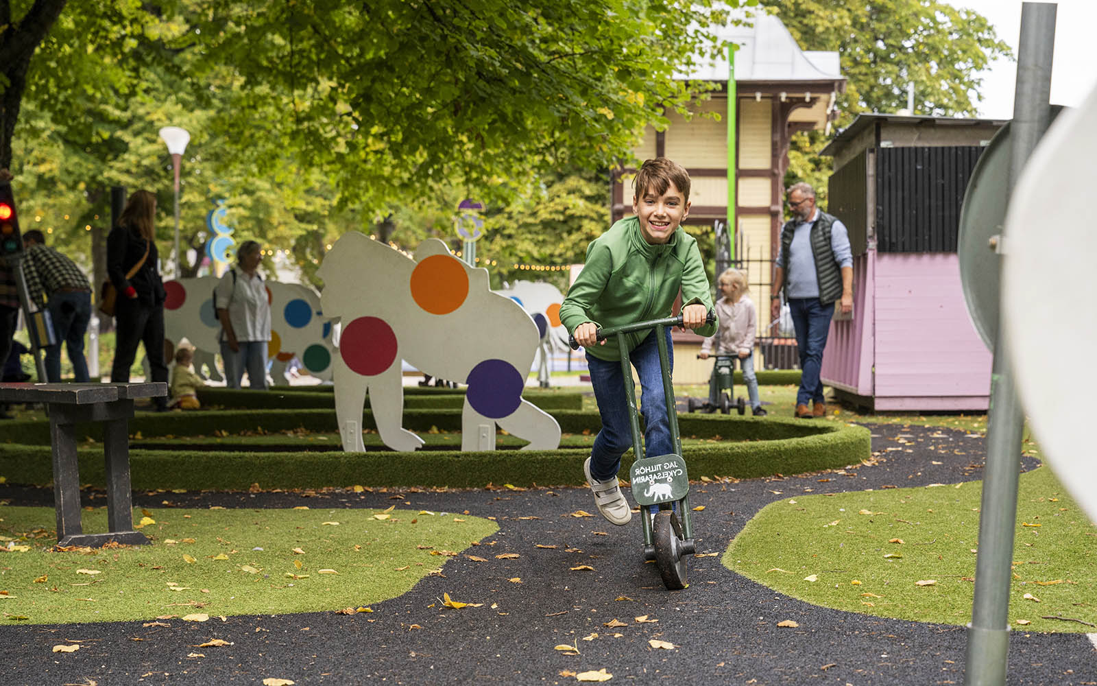 En pojke med brunt hår och en grön tröja åker på en sparkcykel i en lekpark. Bakom ser man en asfalterad stig, gräsmatta och vita trädjur med prickar på.